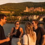 Gruppe Personen auf der Alten Brücke vor Schlosspanorama im Abendlicht trinkt Wein bei einer geführten Weintour Heidelberg